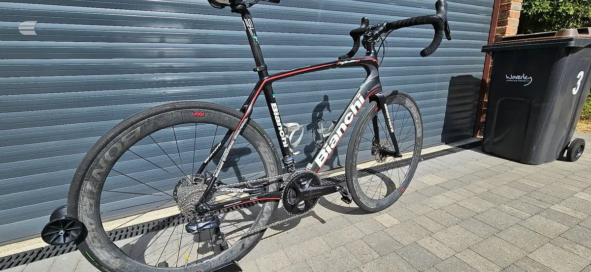 Black and red Bianchi road bike on a stand, drive side, against a blue metal shutter with bins nearby.