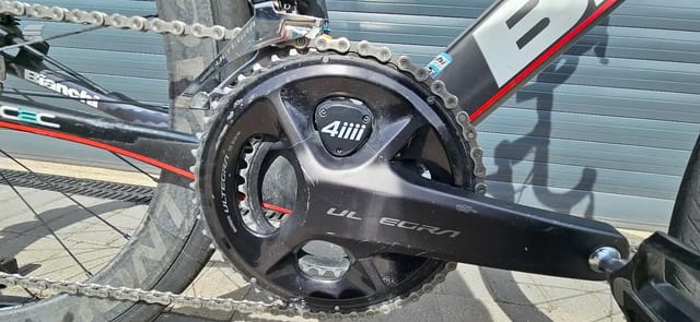 Close-up of black 4iiii crankset and chainring on a black and red road bike, drive side, outdoors against a shutter.