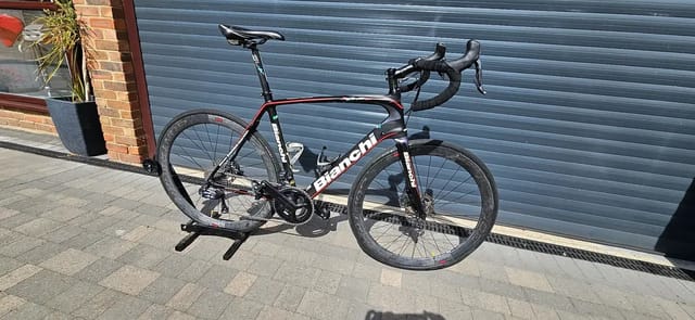Black and red Bianchi road bike on a stand, drive side, against a blue metal shutter with brick building to the left.