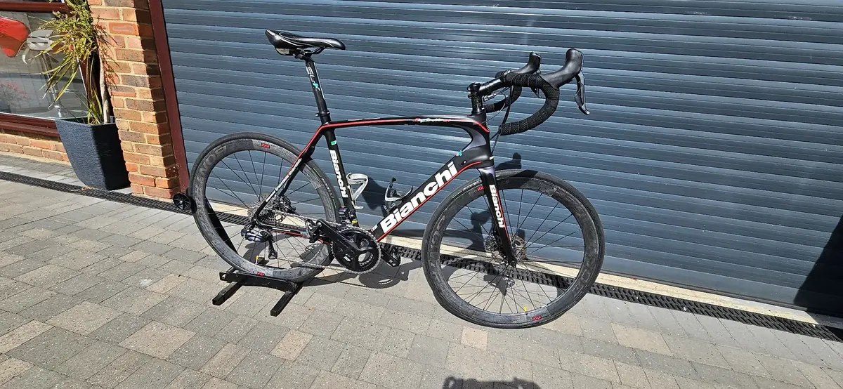 Black and red Bianchi road bike on a stand, drive side, against a blue metal shutter with brick building to the left.