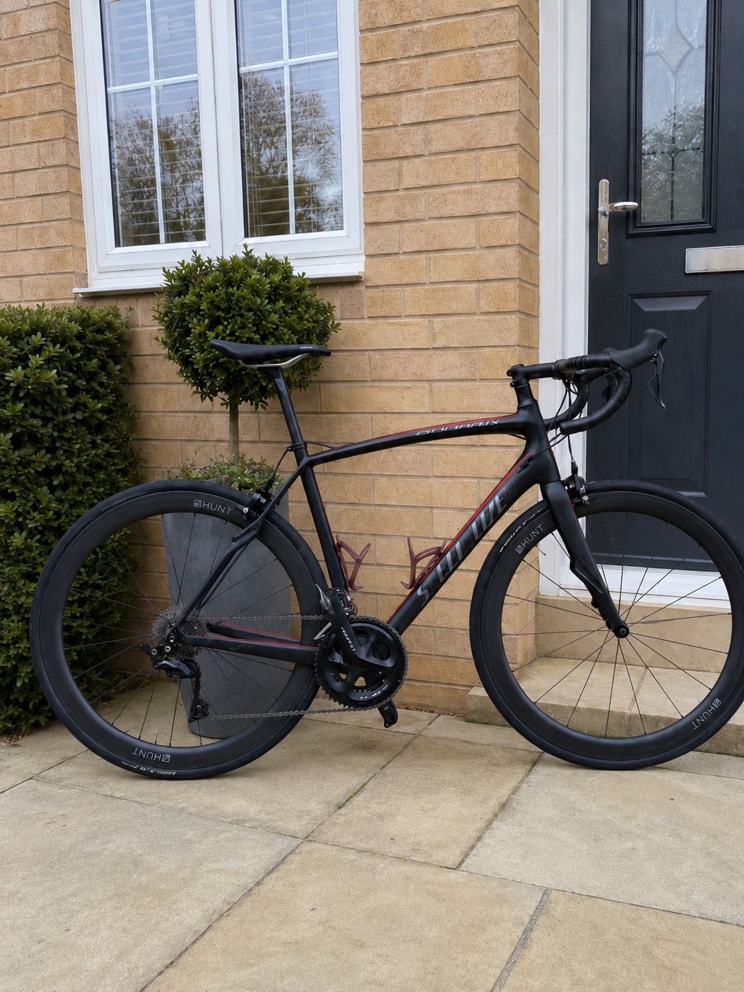 Black and burgundy road bike with drop bars on a paved driveway, drive side, against a brick house with white window and black door.