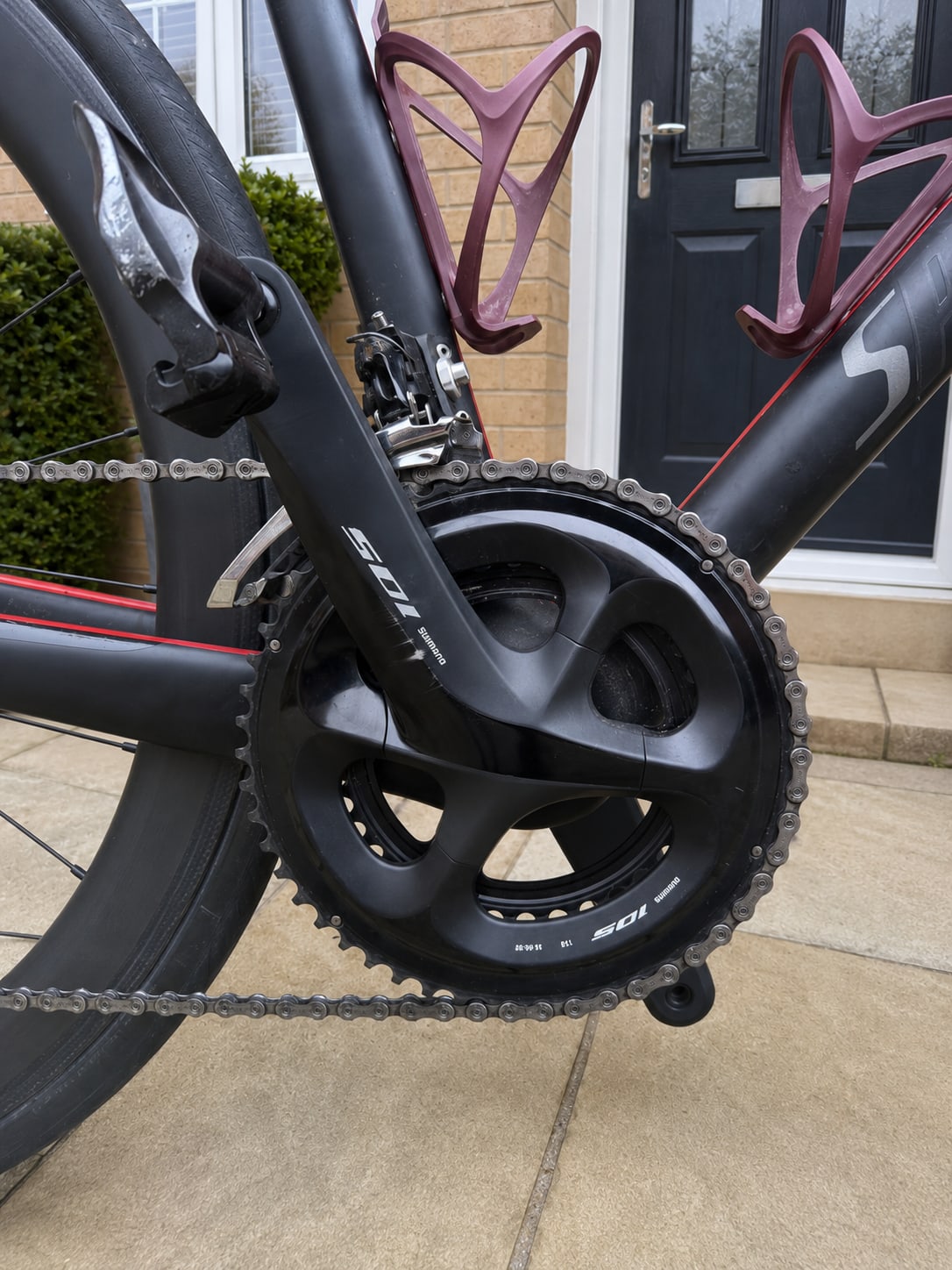 Black road bike crankset and chainring with purple bottle cage, drive side, on a residential driveway.