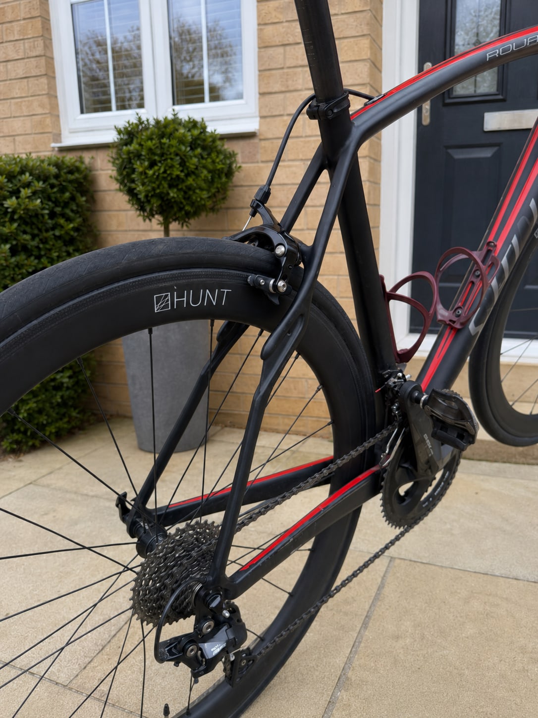 Black and red road bike with HUNT wheels, drive side, on a residential driveway with brick house and topiary in background.