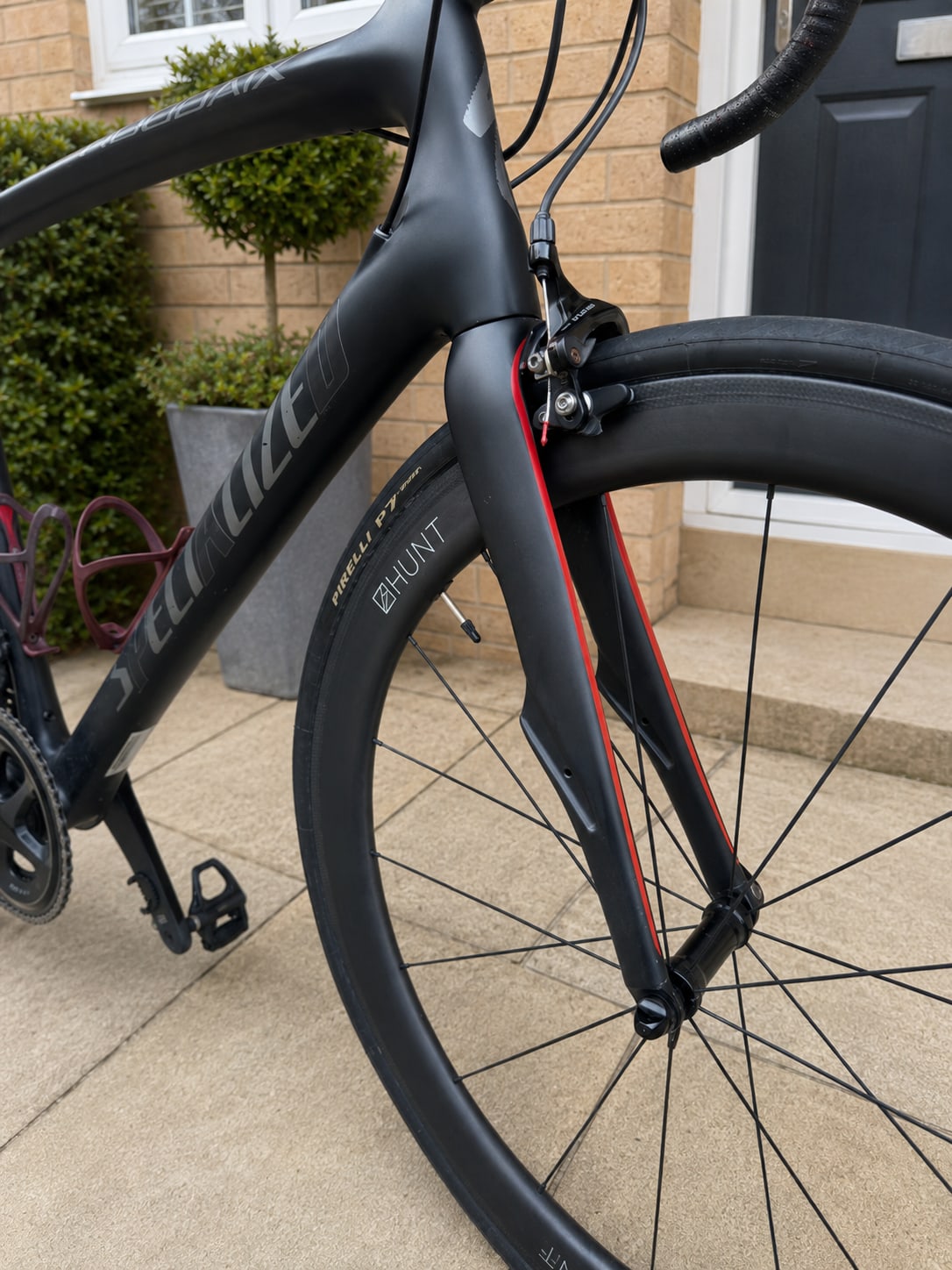 Black road bike front fork and brake detail, drive side, on a residential driveway with brick house and potted plant in background.