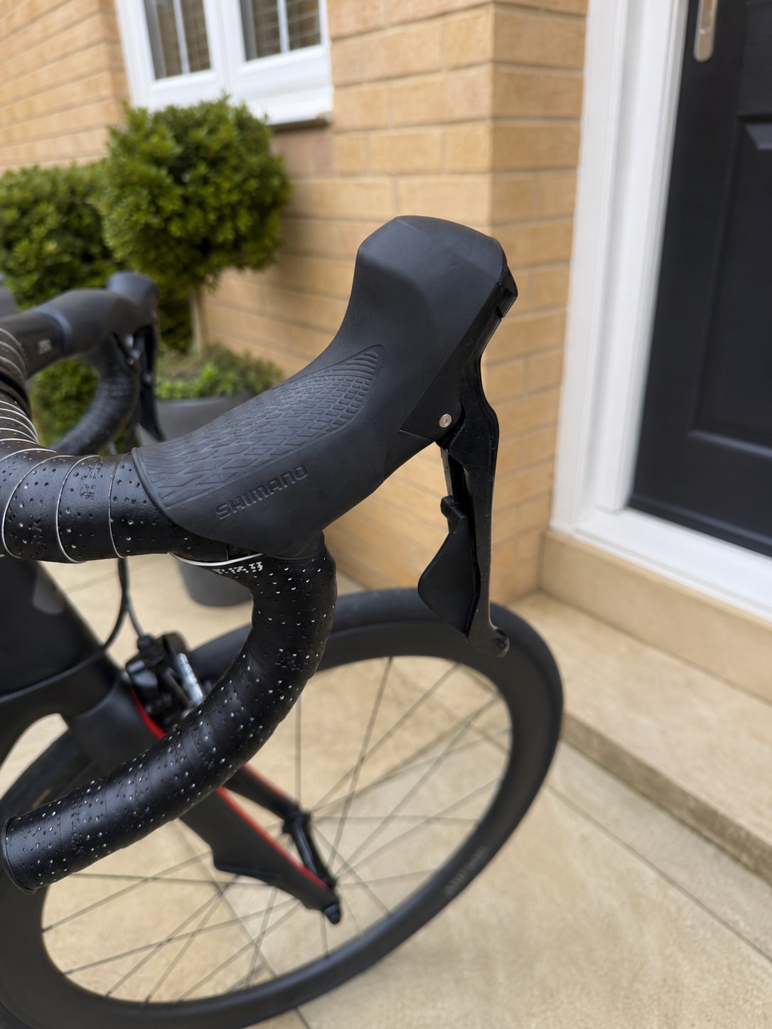 Shimano drop bar road bike shifter and brake lever on black handlebar tape, drive side, against a brick house exterior.