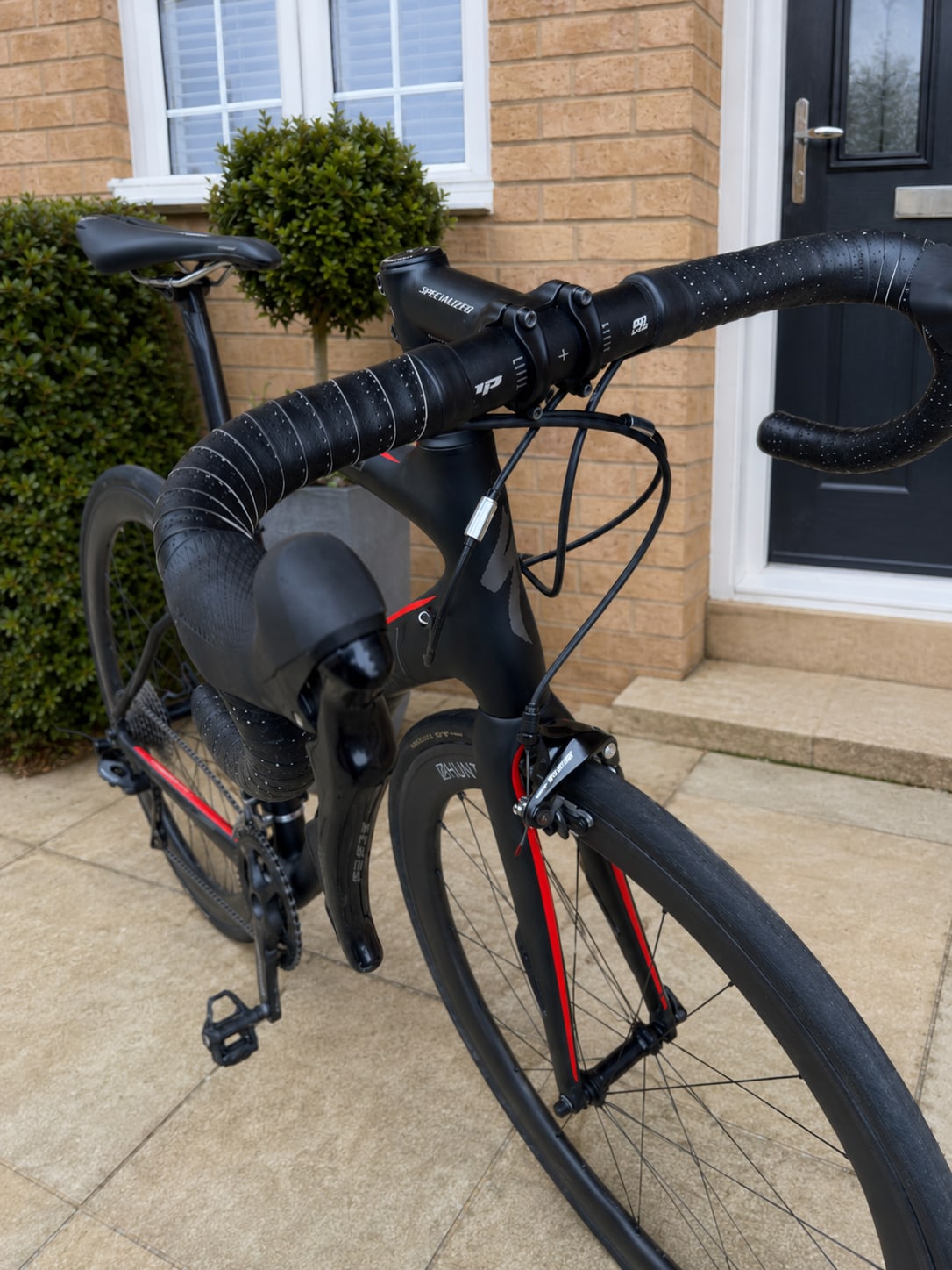 Black and red Specialized road bike on paved driveway, drive side, against brick house with topiary and black door.