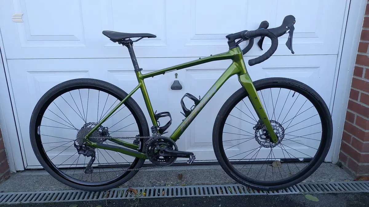 Lime-green gravel bike with disc brakes and drop bars, drive side, leaning against a white garage door with brick wall visible.