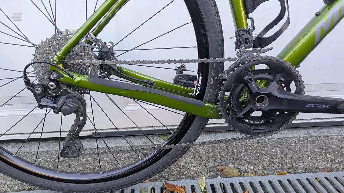Green and black road bike drivetrain detail, drive side, showing crankset, chain, cassette and rear derailleur on concrete ground.