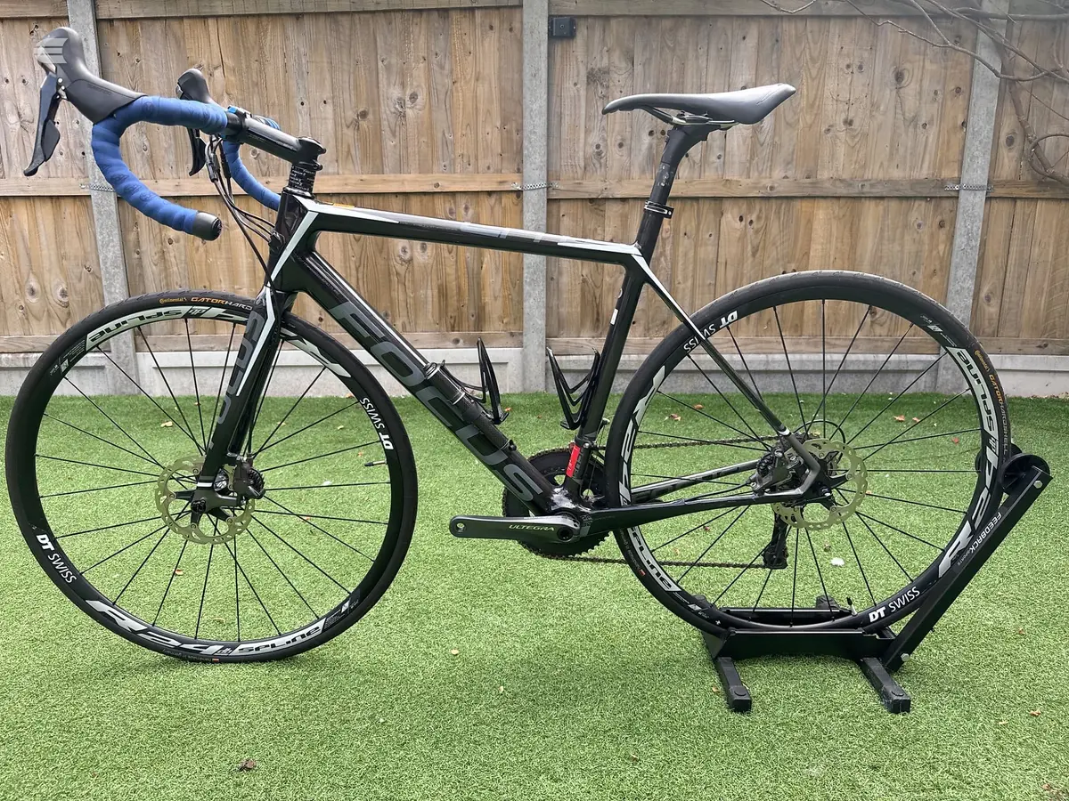 Black road bike with blue drop bars on a stand, drive side view, against a wooden fence and green grass background.