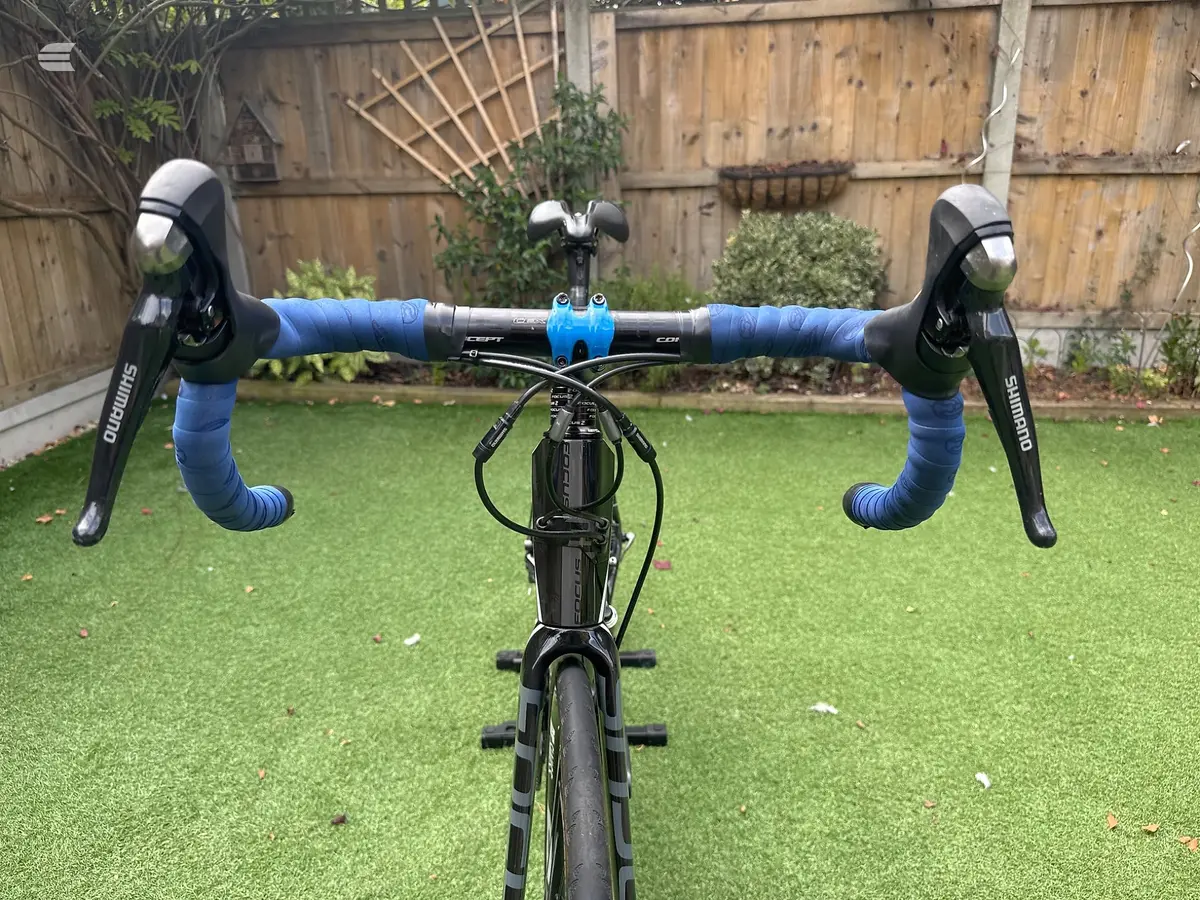 Black road bike with blue drop bars and bar tape, front-on view in a residential garden with wooden fence background.
