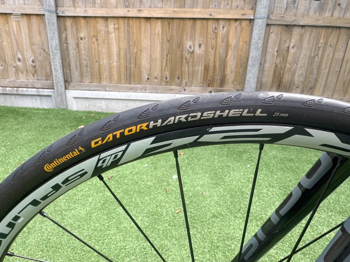 Continental Gator Hardshell 25mm bicycle tyre on a light-coloured rim, angled view against green artificial turf and wooden fence background.