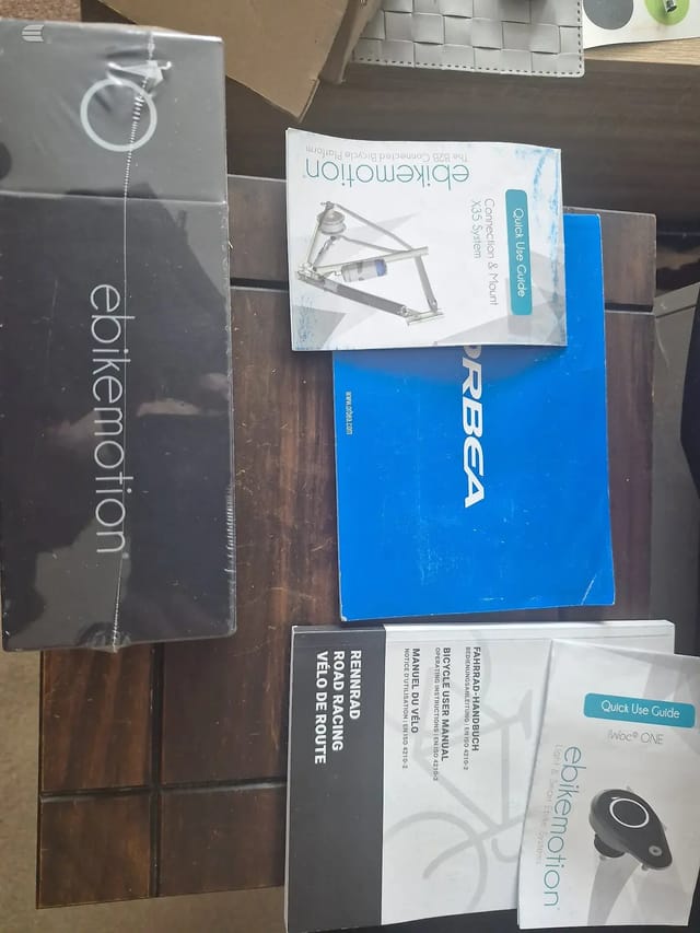 Collection of bicycle product boxes and manuals on a wooden table, including Epikemotion, RBGA, and ebikeemotion branded packaging with quick-use guides.