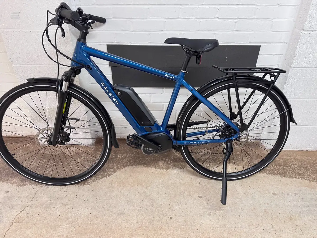 Blue Raleigh Felix electric bike with black mudguards and rear rack, drive side, on a kickstand against a white brick wall.