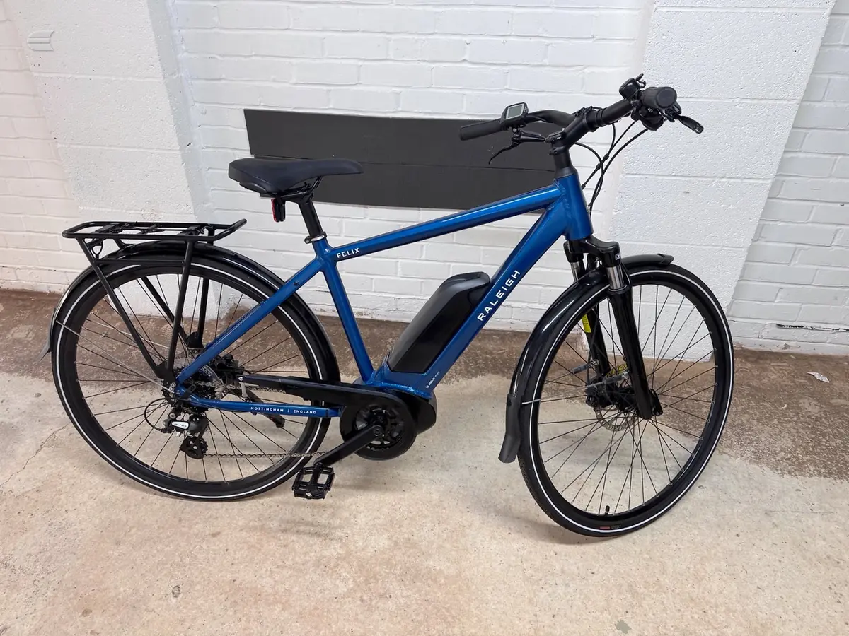 Blue Raleigh Felix electric bike on concrete, drive side, against white brick wall. Black mudguards, front rack, and integrated battery visible.