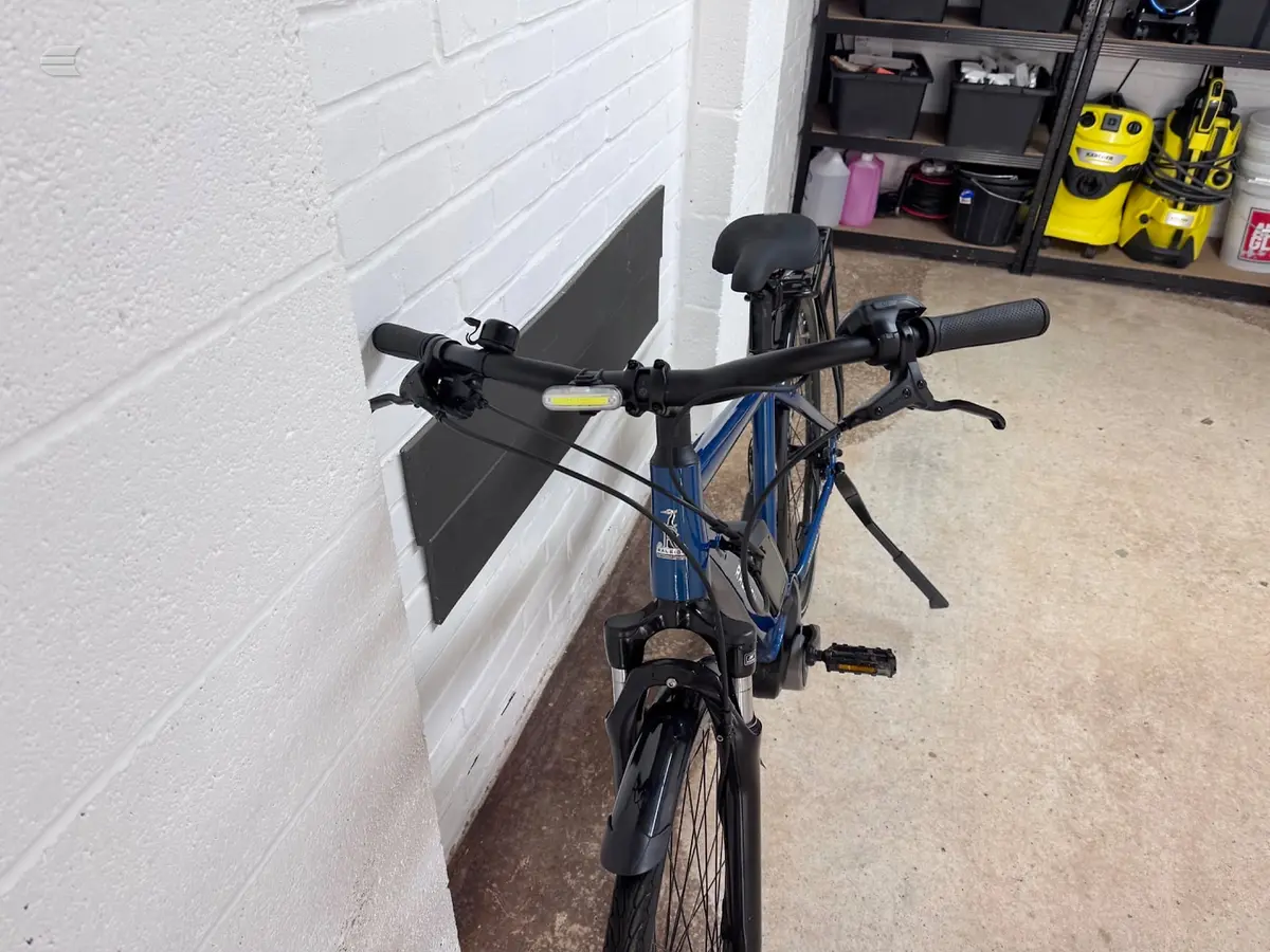 Blue hybrid bike with drop bars, front-facing view in a garage against a white brick wall and garage door, drive side visible.