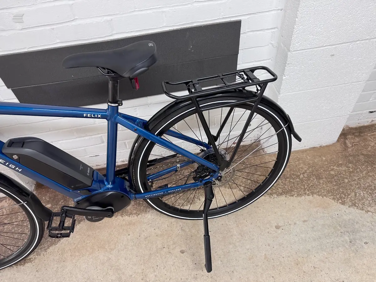 Blue Felix electric bike on kickstand, drive side, against white brick wall and brown ground.