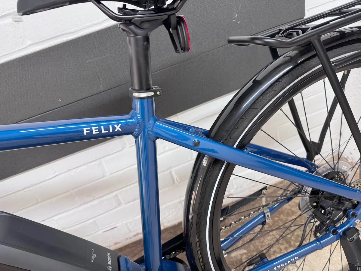 Blue Felix road bike, drive side, leaning against a white brick wall with dark grey panel above.