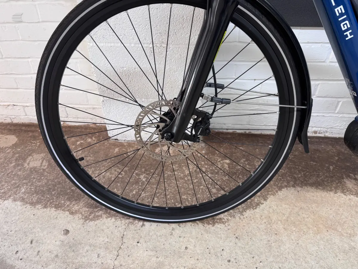 Front wheel of a blue hybrid or gravel bike with disc brake, drive side, against a white brick wall and concrete ground.