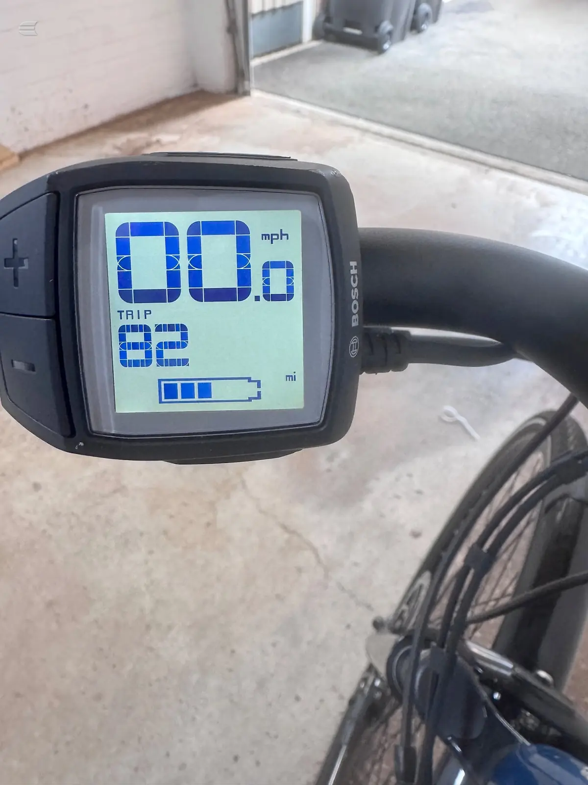 Bosch bicycle computer display mounted on handlebars, showing speed of 00.0 mph, trip distance of 82 miles, and battery indicator, outdoors on concrete