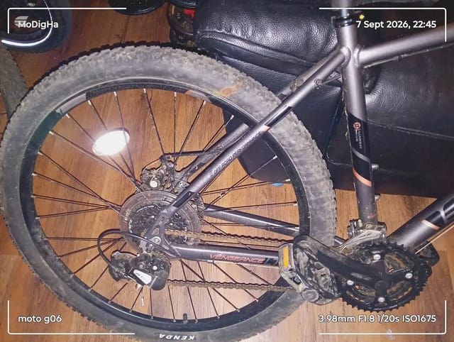 Close-up of rear wheel and drivetrain on a black mountain bike, drive side, indoors on wooden floor with black saddle bag visible.