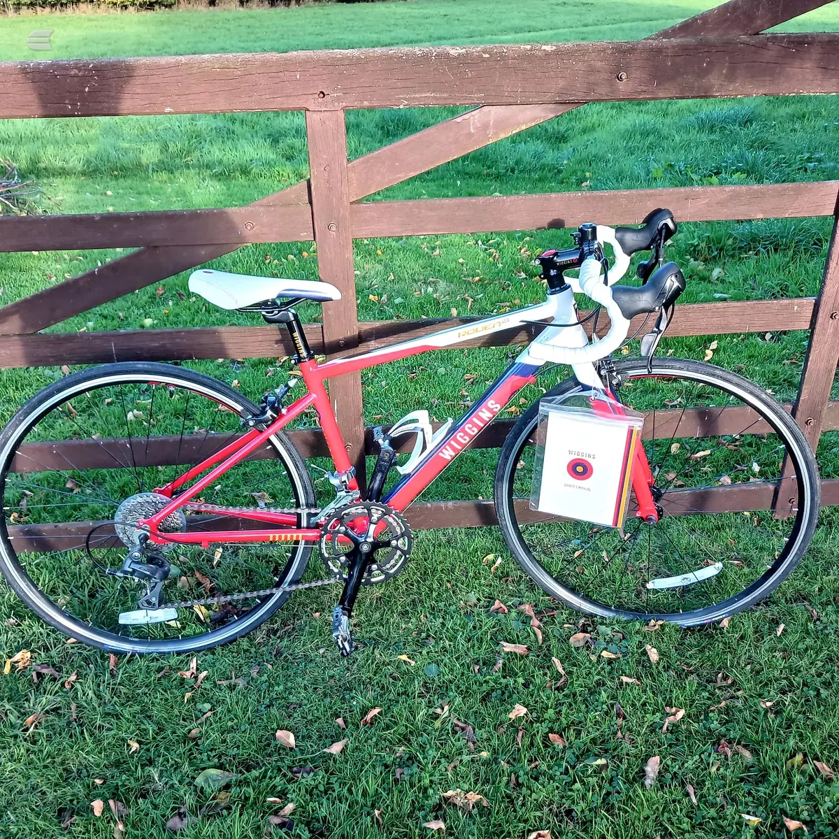 Red and white Niner road bike with drop bars, drive side, leaning against a wooden ranch fence with green grass and fallen leaves in background.
