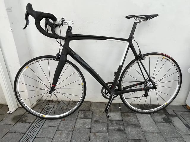 Black Avanti road bike, drive side, leaning against white wall on paved ground.