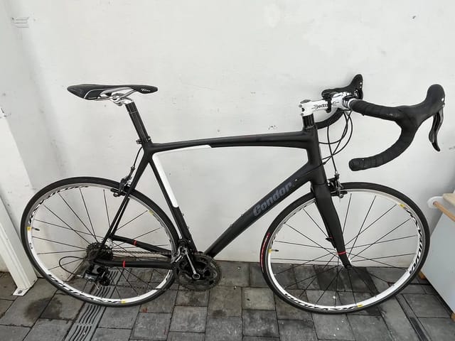 Black Condor road bike with white stem and black drop bars, drive side, leaning against white wall on paved ground.