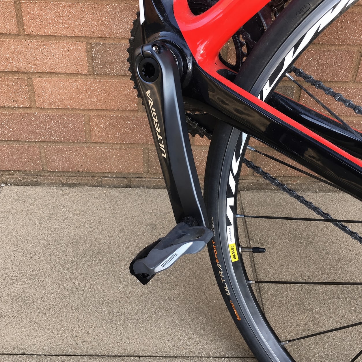 Close-up of Shimano Ultegra derailleur and chain on red and black road bike, drive side, leaning against brick wall.
