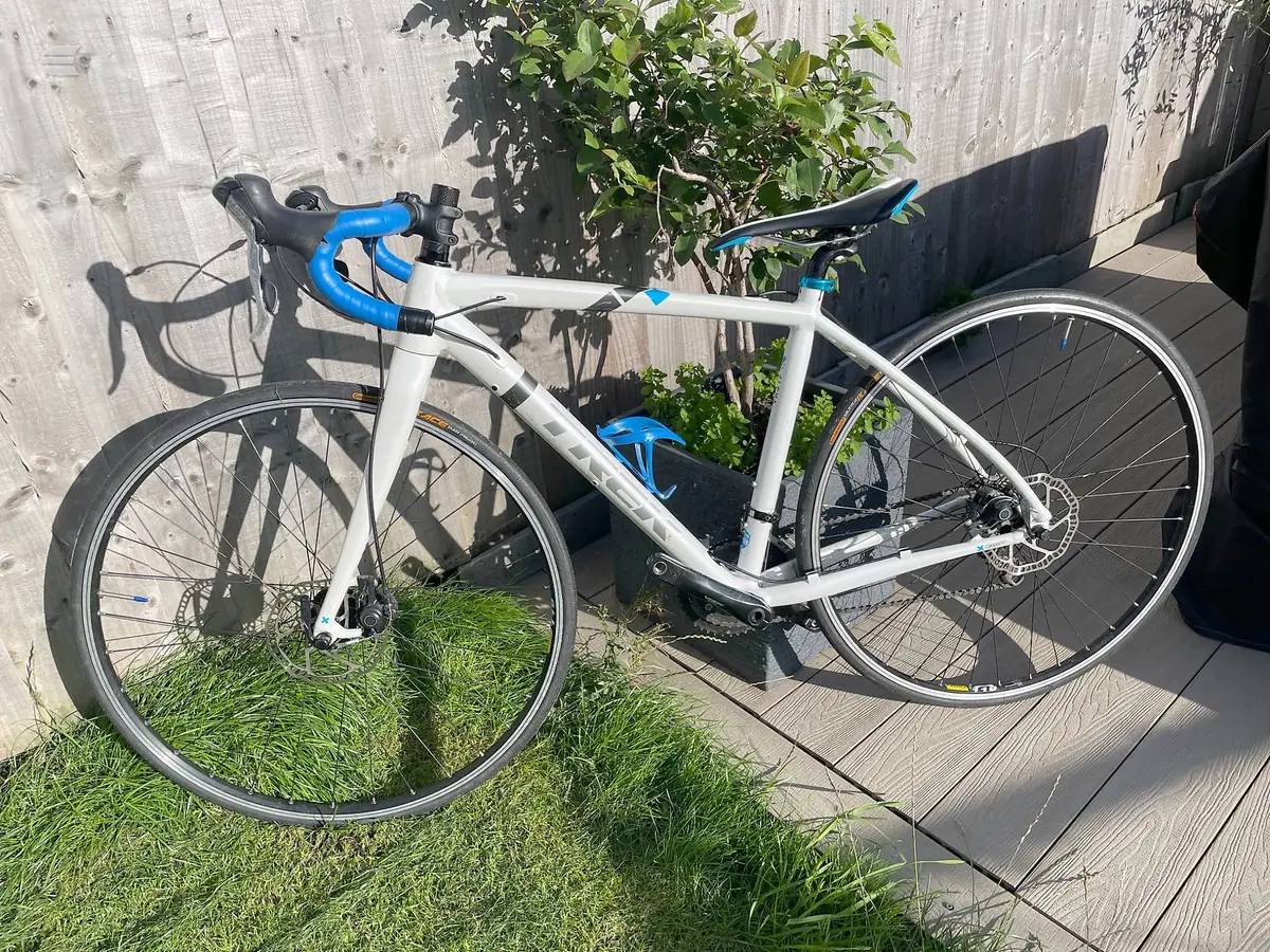 White and blue gravel bike with drop bars, drive side, on a wooden deck against a weathered fence with potted plants.