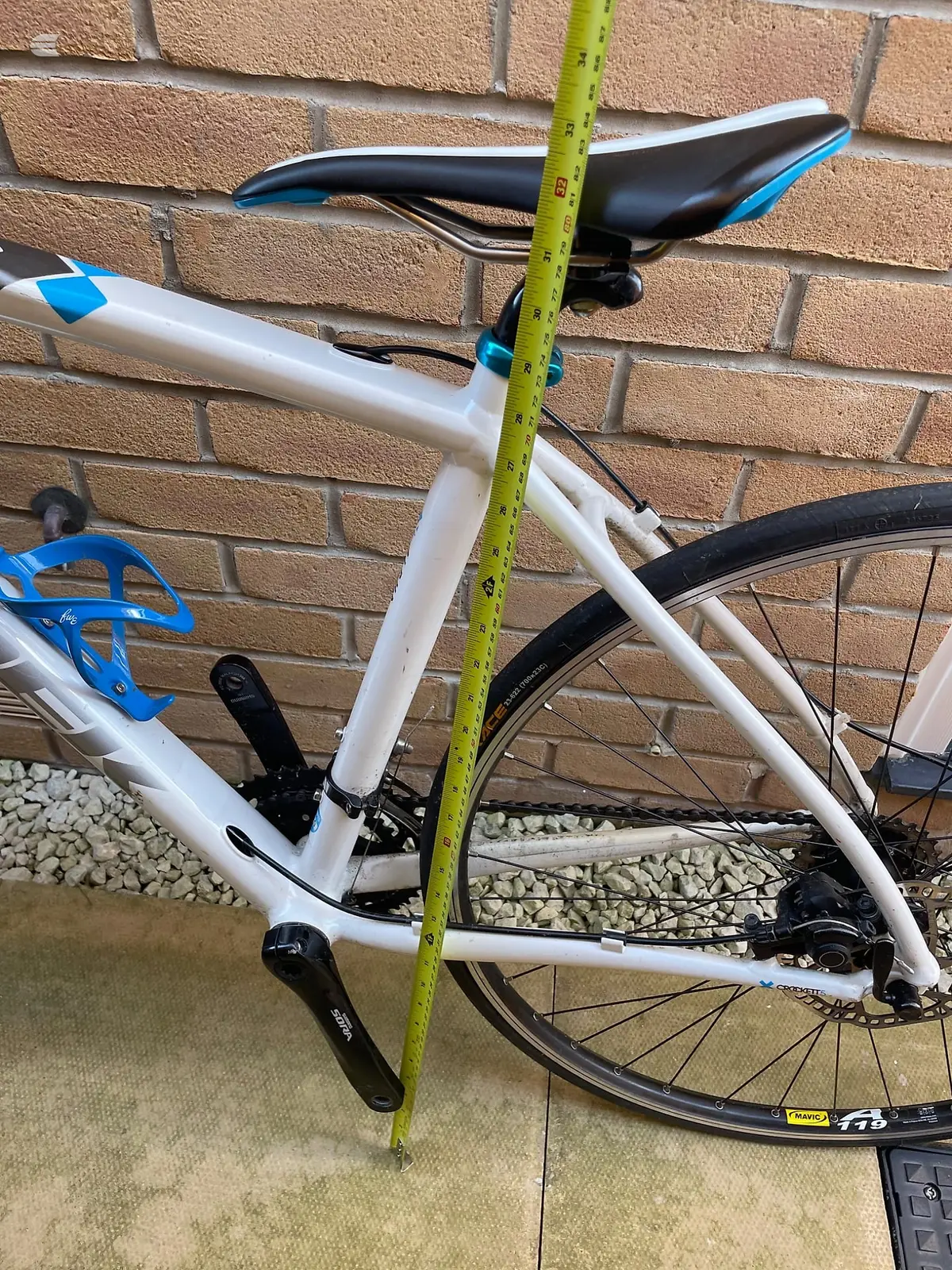 White and blue hybrid bike with black saddle and components, non-drive side, leaning against brick wall with yellow measuring tape across frame.