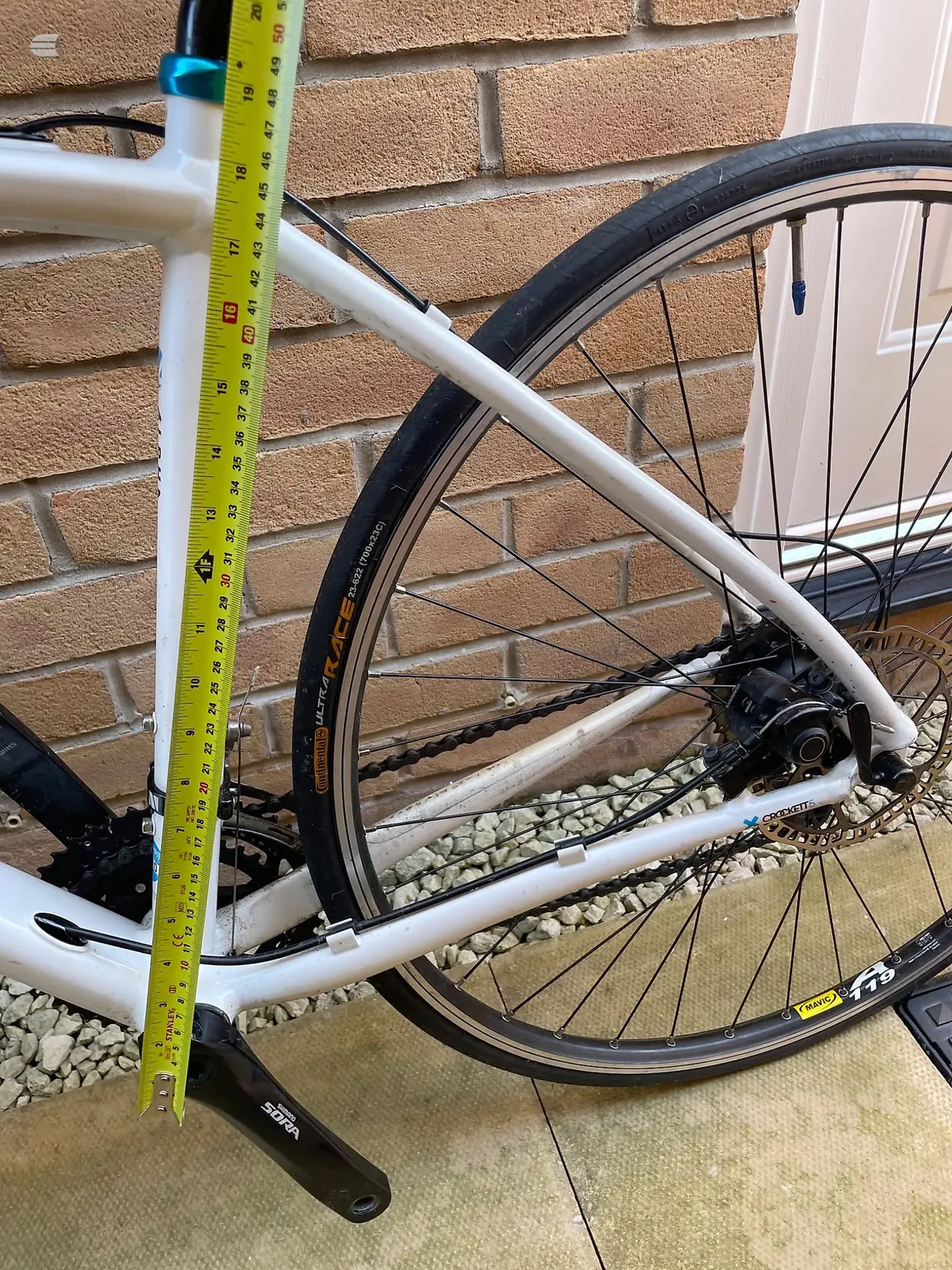 White hybrid bike on a stand against a brick wall, non-drive side, with a yellow tape measure held against the frame for scale.