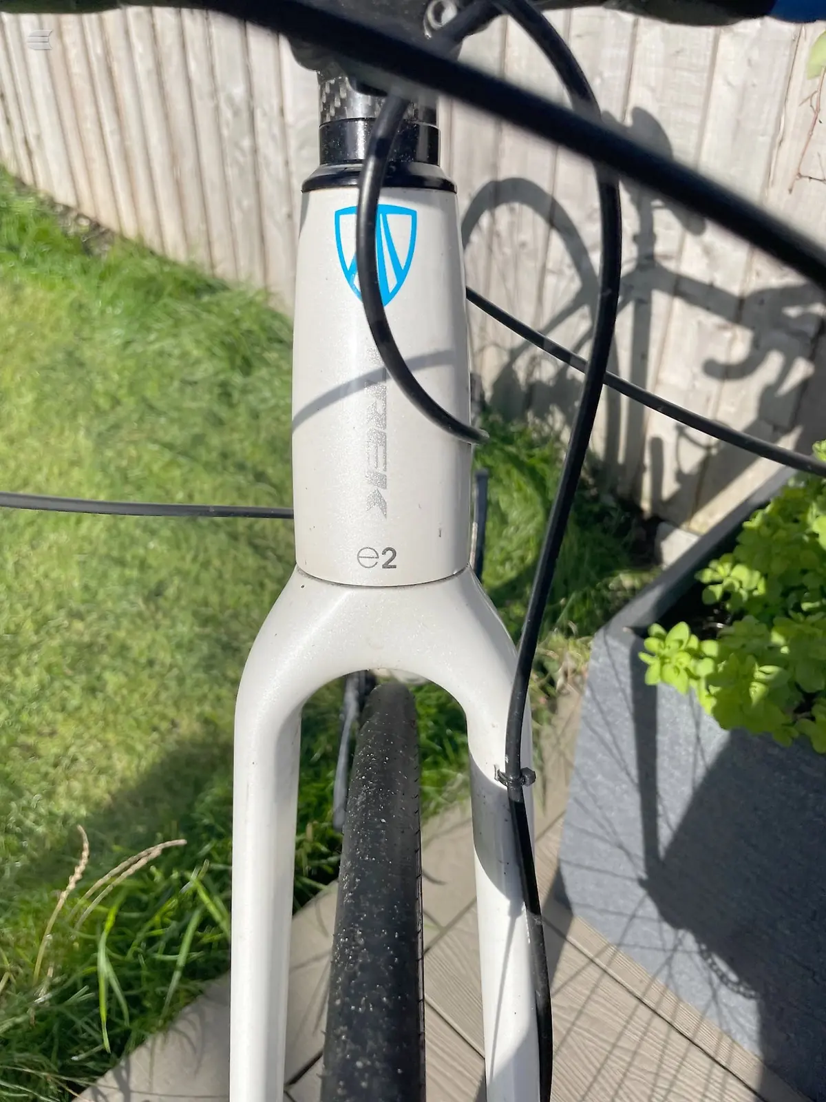 White e2 gravel bike frame with blue shield logo, non-drive side, outdoors against a metal fence with grass and patio visible.