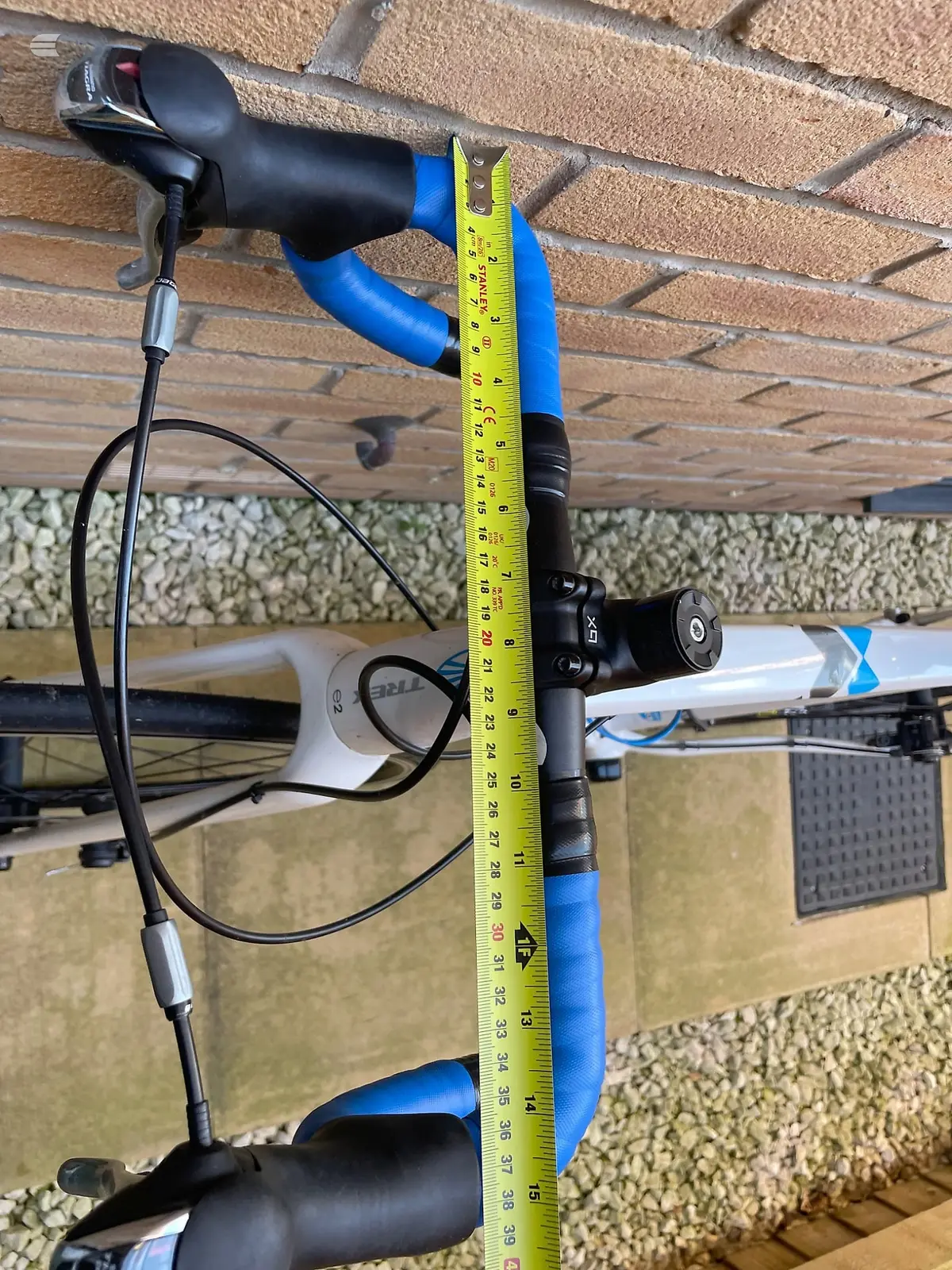 Blue and black electric scooter handlebar and stem assembly measured against a tape measure, mounted on brick wall background.