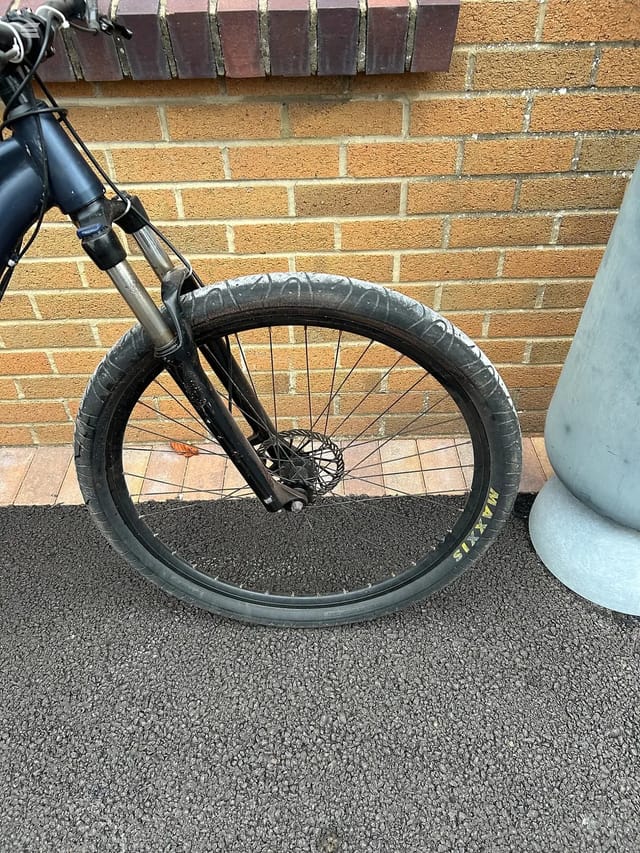 Black gravel bike front wheel with disc brake, close-up against brick wall and tarmac, non-drive side.