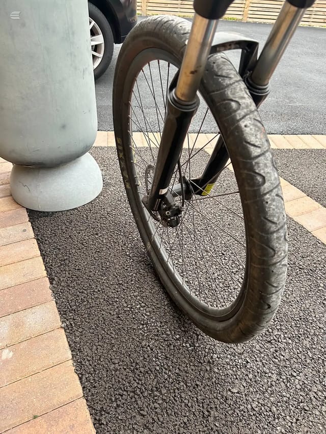 Front wheel and fork of a gravel or hybrid bike with disc brakes, parked against a white bollard on pavement, non-drive side view.