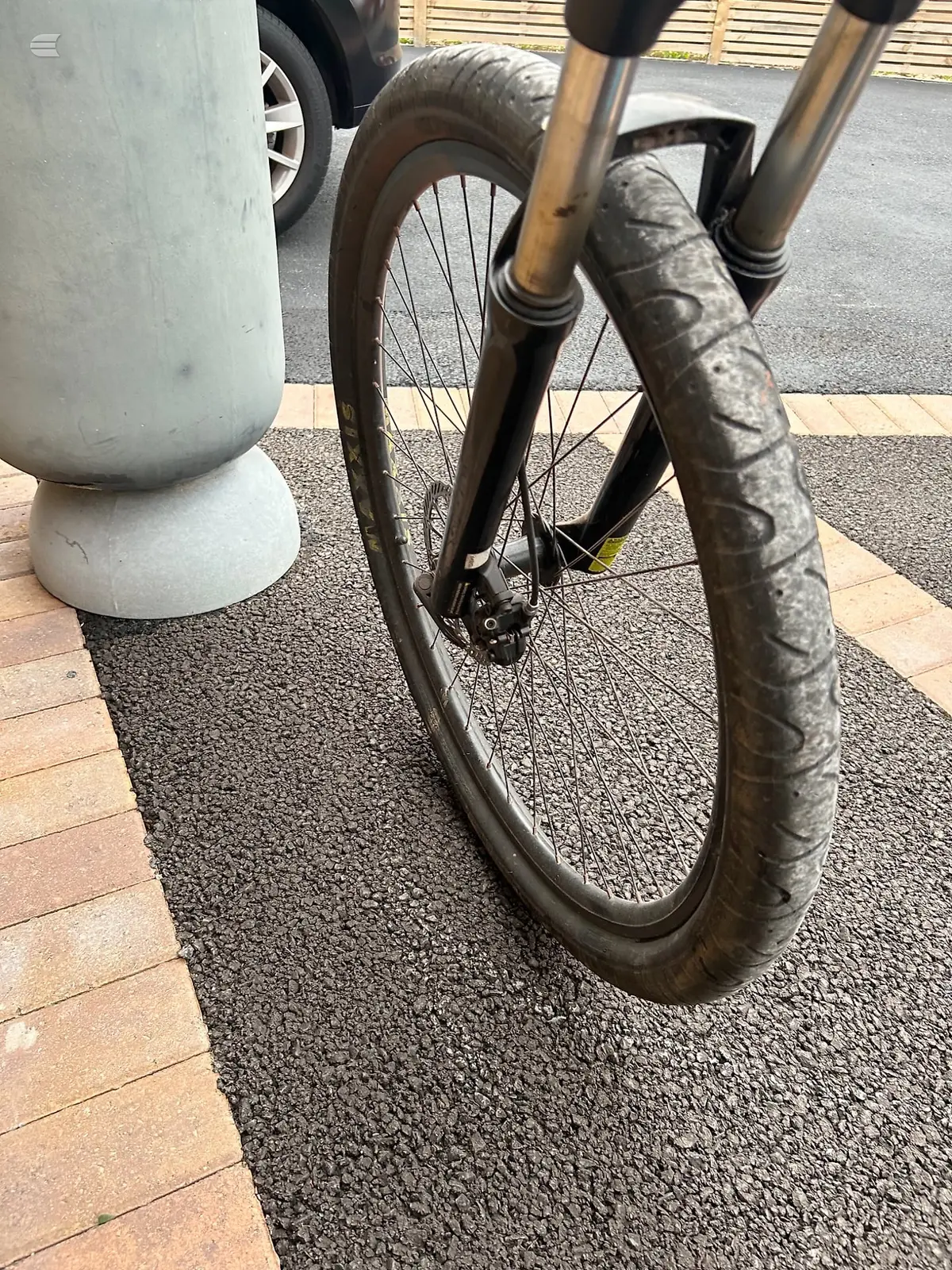 Front wheel and fork of a gravel or hybrid bike with disc brakes, parked against a white bollard on pavement, non-drive side view.