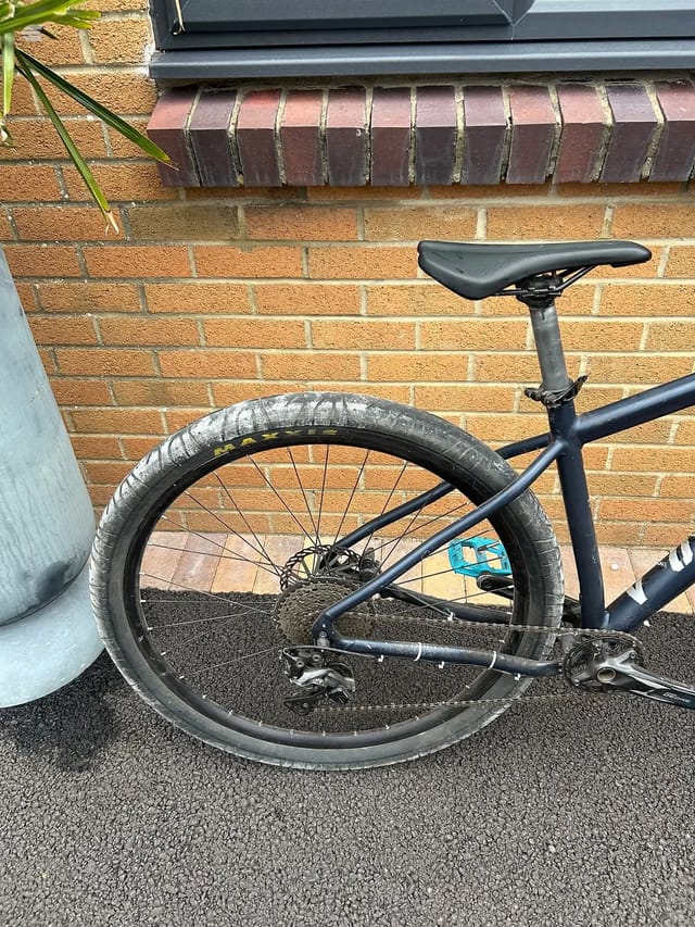 Black gravel bike rear wheel and drivetrain, drive side, leaning against brick wall with dark window above.