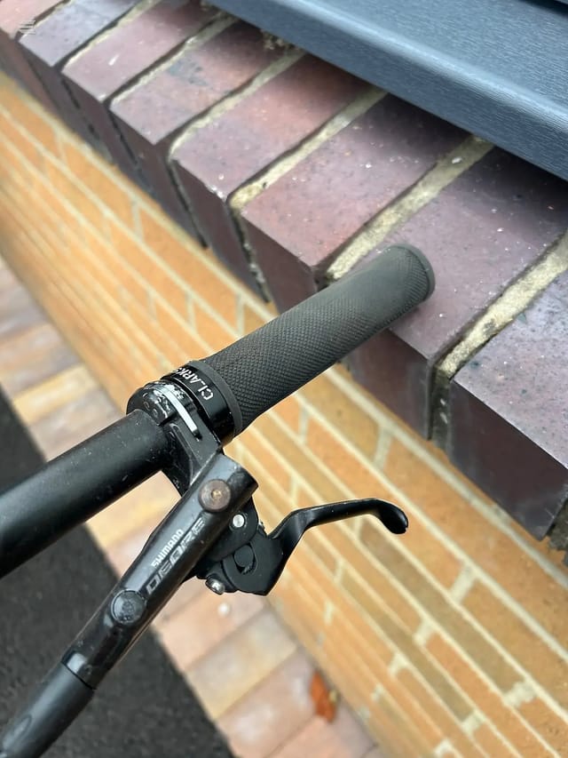 Close-up of bicycle handlebars and brake lever, black grips and Shimano components, against brick building and metal steps.
