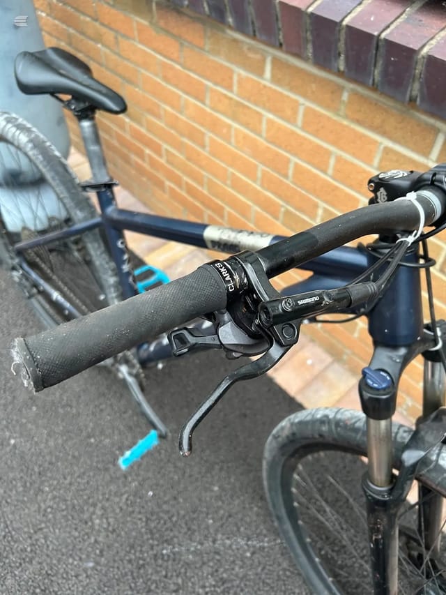 Close-up of black drop handlebars and Shimano brake levers on a hybrid or gravel bike, against a brick wall background.