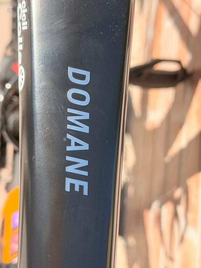 Close-up of black bicycle down tube with blue 'DOMANE' text decal, non-drive side, outdoors in sunlight.