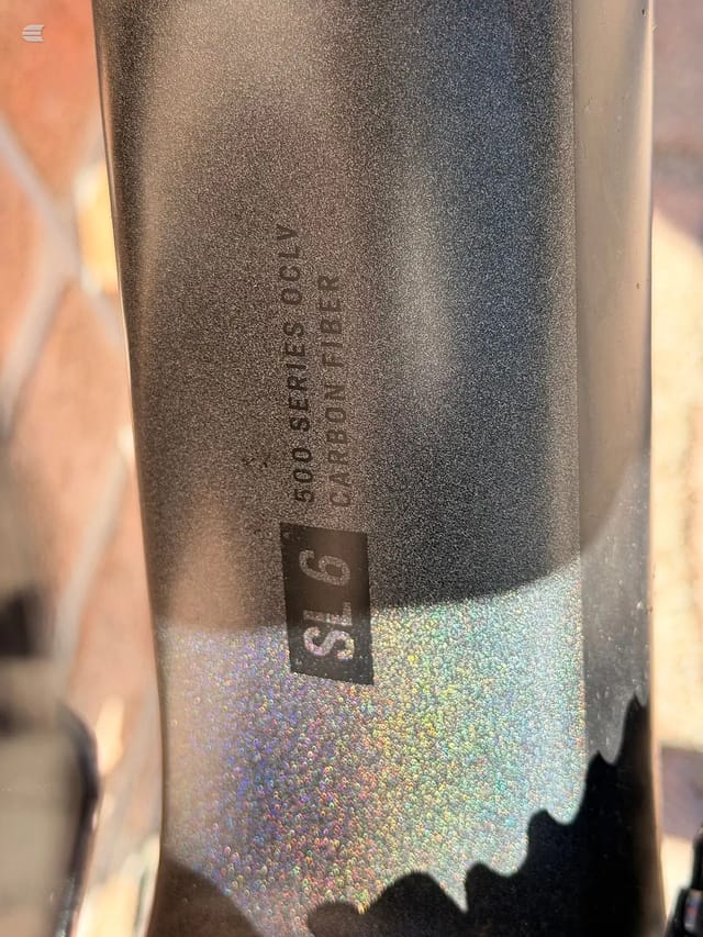 Close-up of grey metallic bicycle seatpost with embossed branding text and holographic sticker detail, angled against blurred background.
