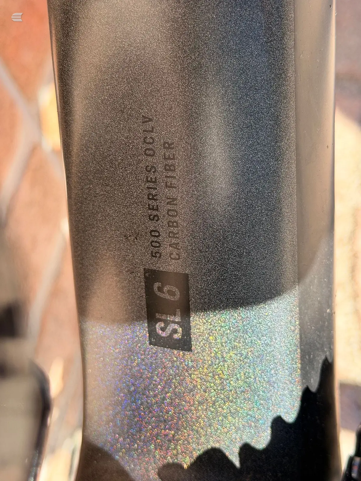 Close-up of grey metallic bicycle seatpost with embossed branding text and holographic sticker detail, angled against blurred background.