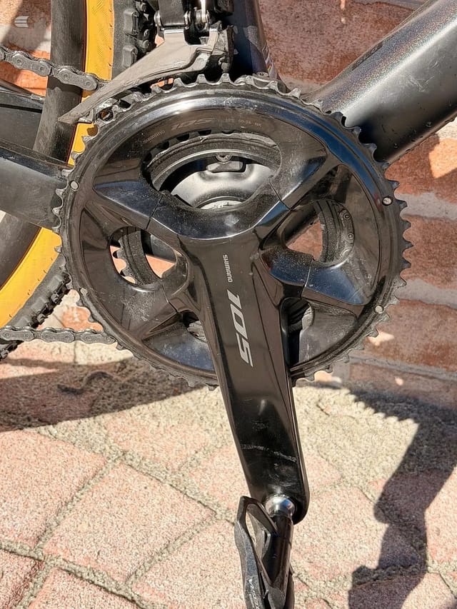Shimano 105 crankset and chainrings on a road bike, drive side, close-up on pavement with yellow and grey bike frame visible.