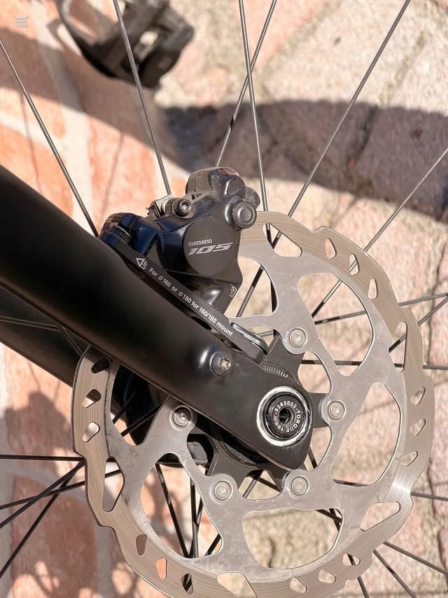 Shimano 105 hydraulic disc brake caliper and rotor on bicycle frame, close-up detail shot against brick background.