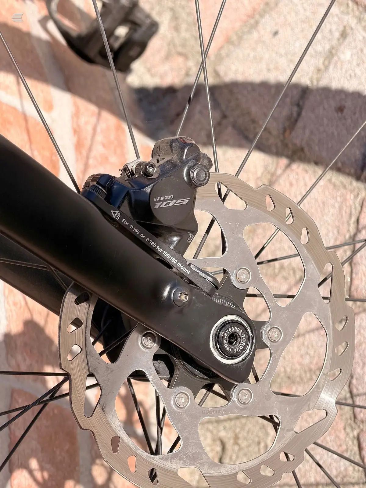Shimano 105 hydraulic disc brake caliper and rotor on bicycle frame, close-up detail shot against brick background.