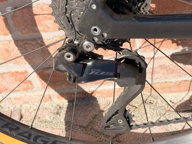 Shimano 105 rear derailleur on yellow bike, drive side close-up against brick pavement.
