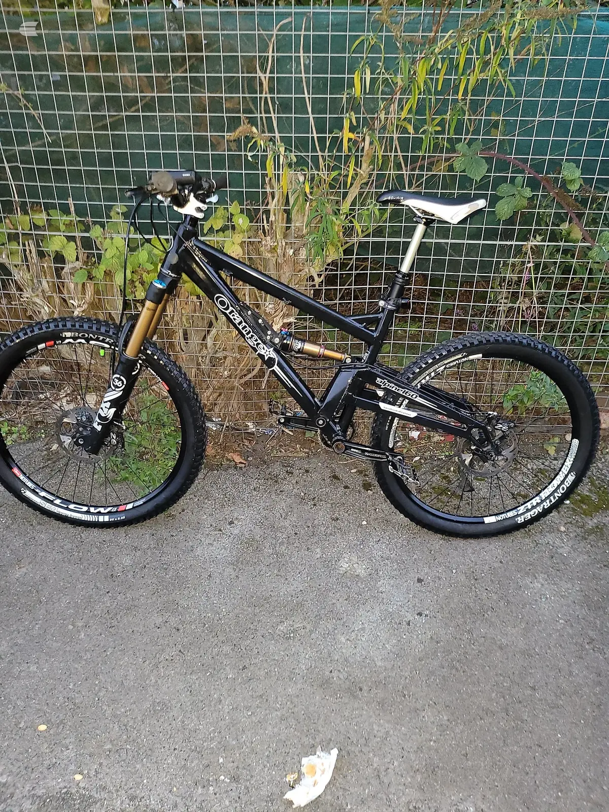 Black full-suspension mountain bike, drive side, on asphalt against a green mesh fence with dried plants.