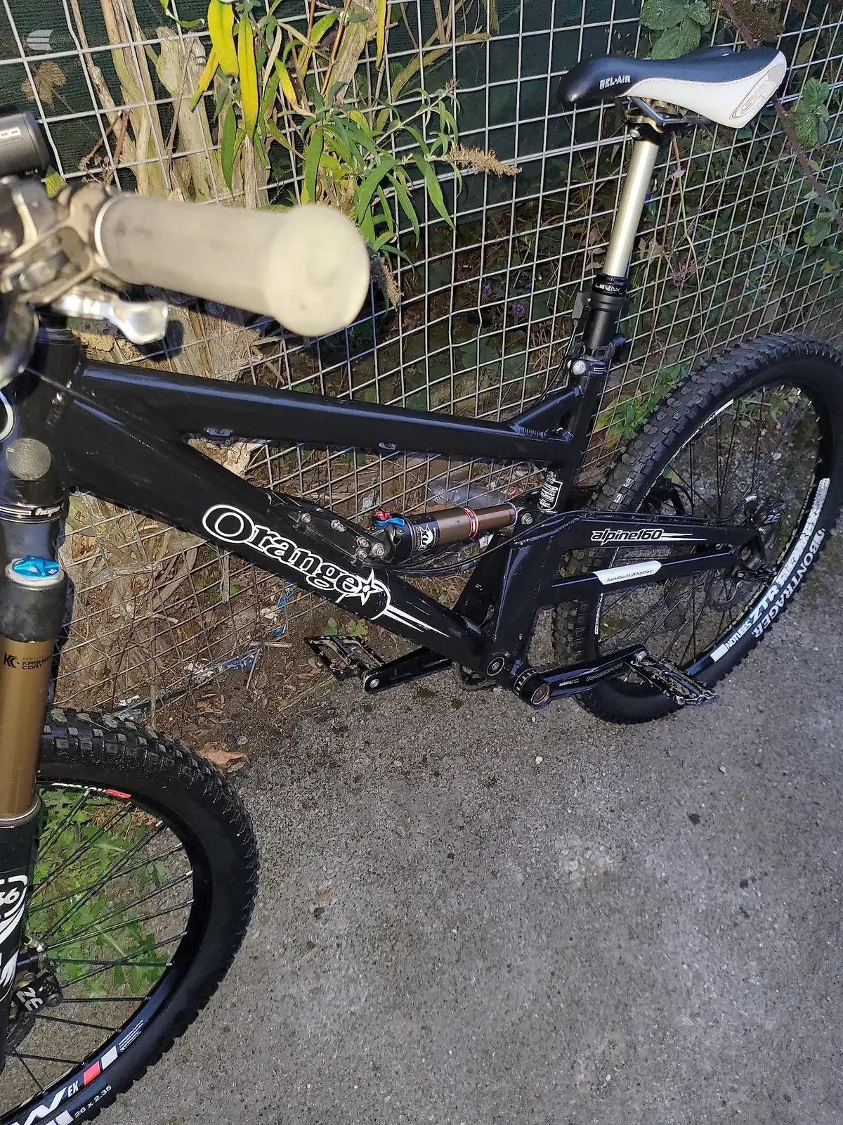 Black Orange full-suspension mountain bike, drive side, leaning against wire fence in outdoor setting with dried plants and vegetation.