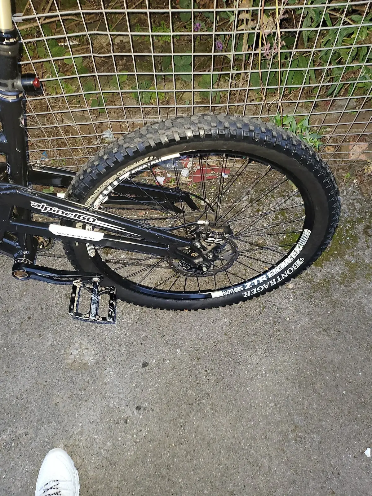 Black Diamondback mountain bike rear wheel with knobby tyre, non-drive side, on pavement against wire fence and garden background.