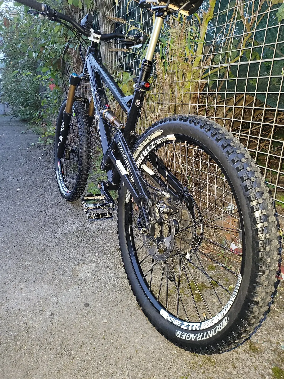 Black hardtail mountain bike, rear wheel close-up, drive side, leaning against wire fence in garden setting.